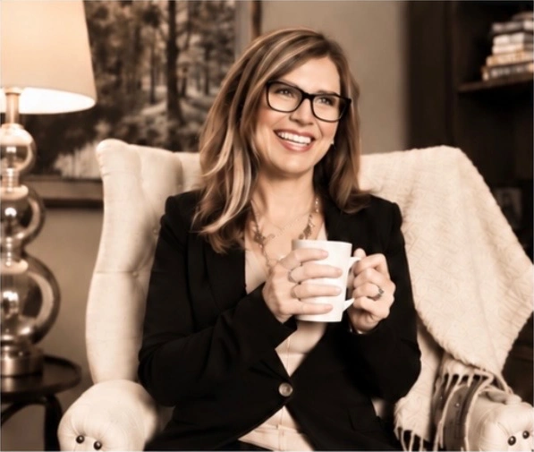 A smiling woman in glasses holding a white mug while seated in a welcoming therapy office